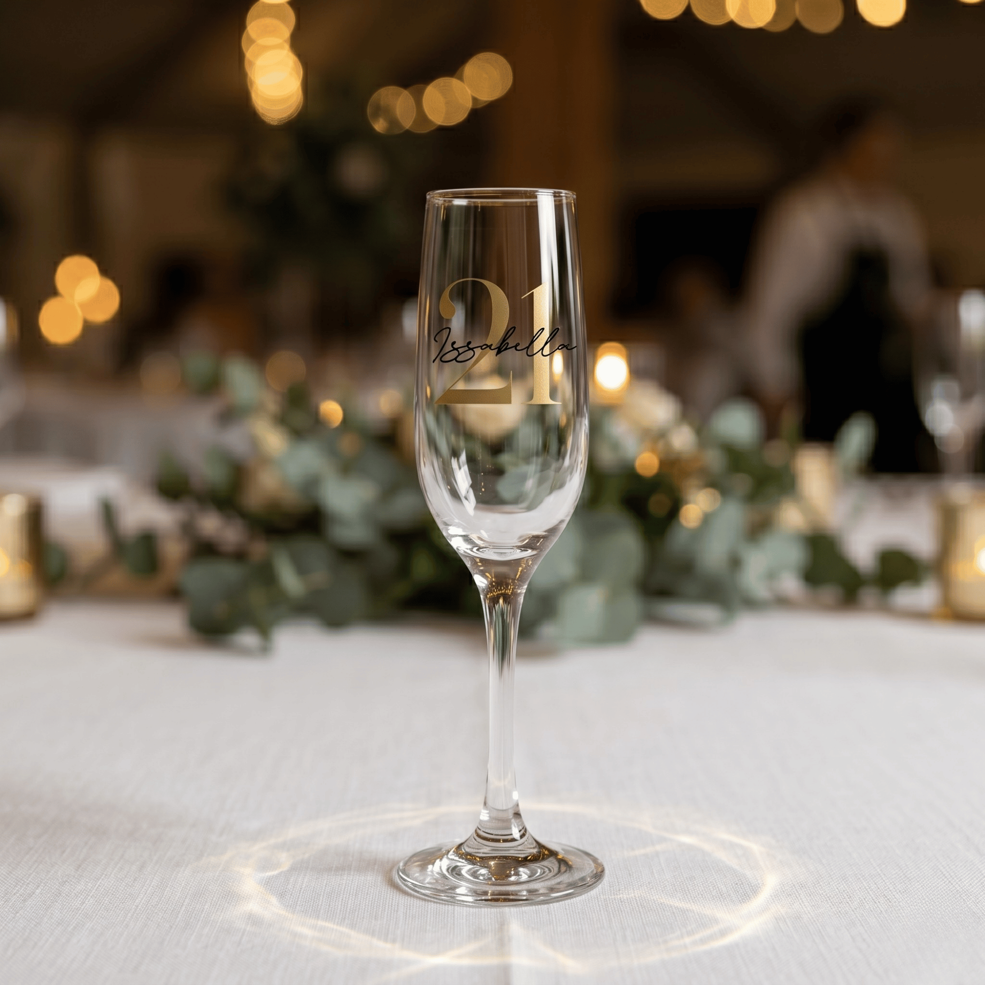 Champagne flute with engraved number 21 on a table with decorative lights and greenery