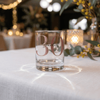 Clear glass with '30 Scarlett' on a table with decorative lights and flowers