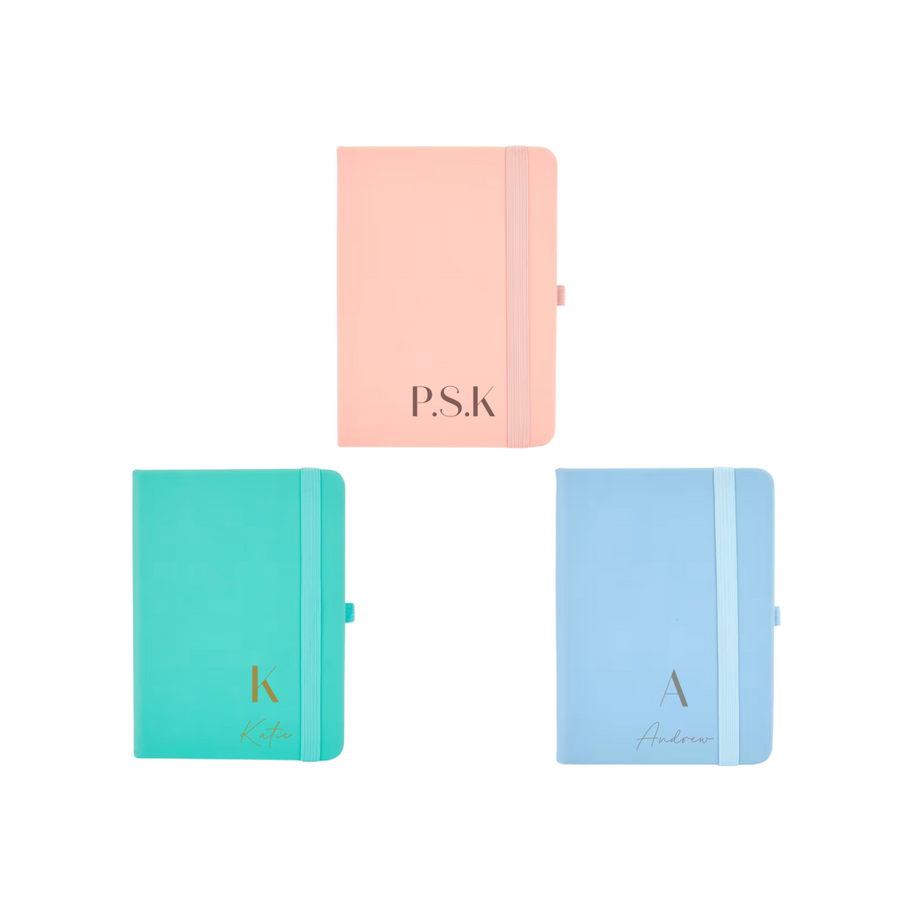 Flat lay of three personalised A6 vegan leather notebooks in pastel peach, teal, and blue colours with custom initials and names on covers.