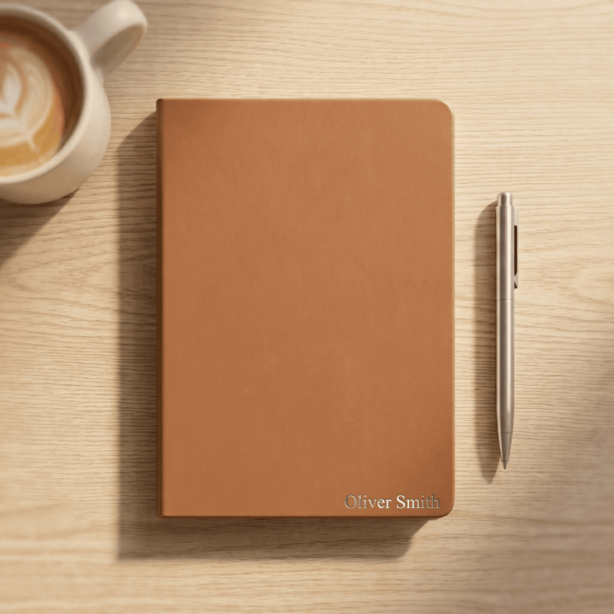 A tan leather notebook with 'Oliver Smith' personalized at the bottom right, styled on a light wood tabletop next to a silver pen and a cup of coffee.