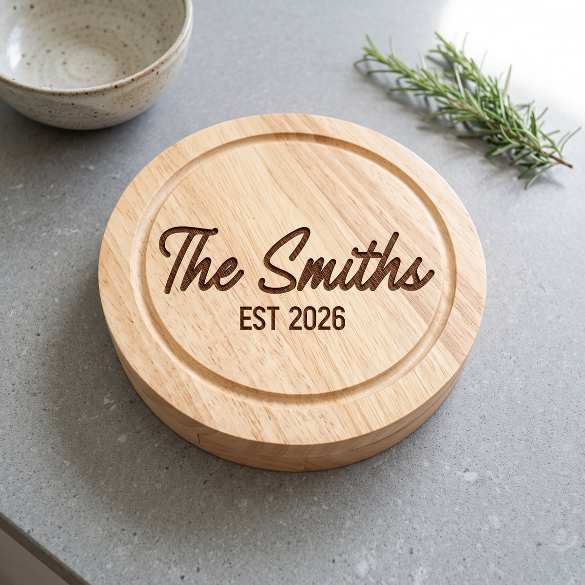 A round natural wood cheese board or trivet engraved with 'The Smiths EST 2026' in dark brown lettering, placed on a gray countertop with a ceramic bowl and rosemary sprig in the background.