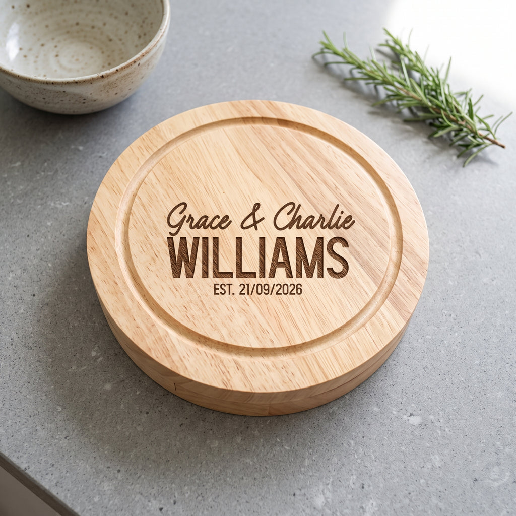 A round natural wood cutting board with engraved personalised text reading 'Grace & Charlie WILLIAMS EST. 21/09/2026', shown on a gray countertop with a ceramic bowl and rosemary sprig nearby.
