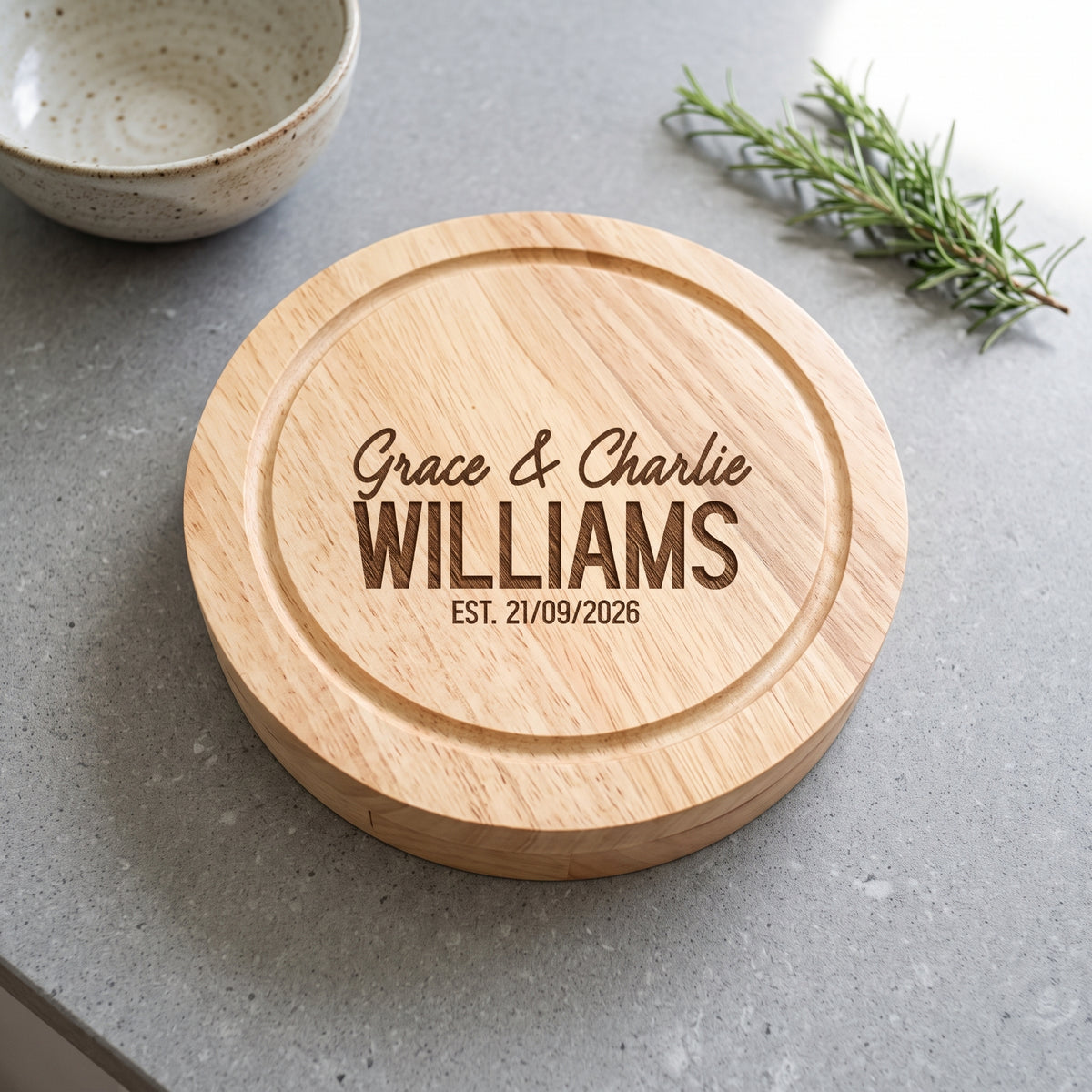 A round natural wood cutting board with engraved personalised text reading 'Grace & Charlie WILLIAMS EST. 21/09/2026', shown on a gray countertop with a ceramic bowl and rosemary sprig nearby.