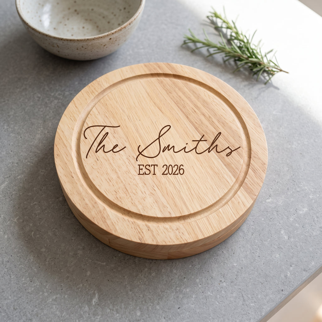 A round natural wood serving board engraved with 'The Smiths EST 2026' in dark brown script and block lettering, placed on a gray stone surface with a ceramic bowl and rosemary sprig in the background.