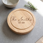 A round natural wood serving board engraved with 'The Smiths EST 2026' in dark brown script and block lettering, placed on a gray stone surface with a ceramic bowl and rosemary sprig in the background.