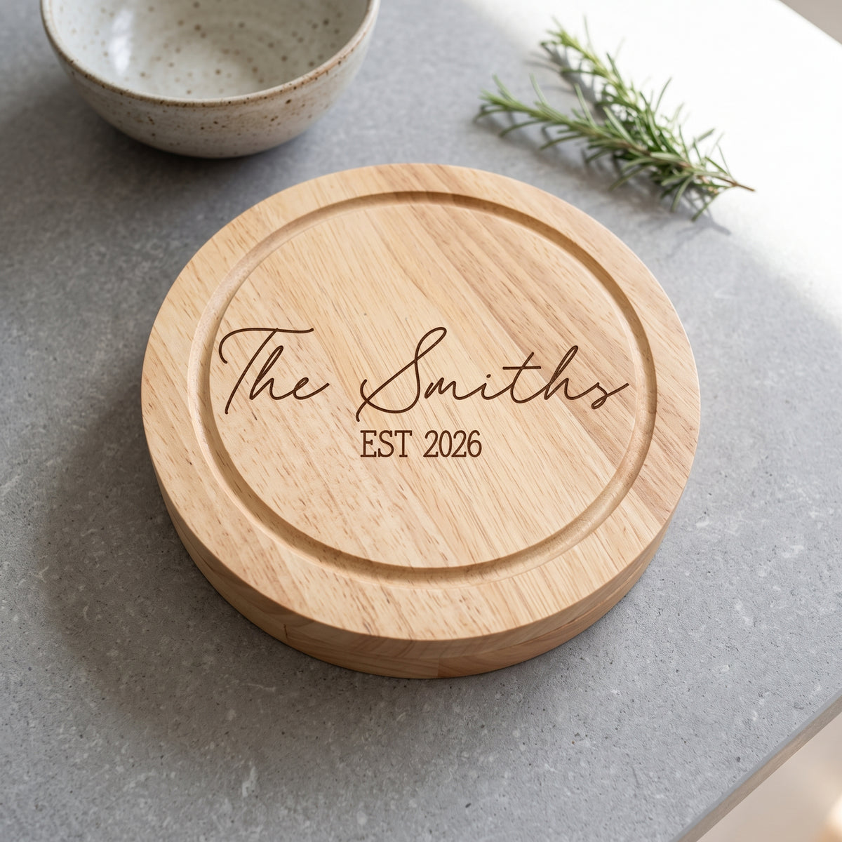 A round natural wood serving board engraved with 'The Smiths EST 2026' in dark brown script and block lettering, placed on a gray stone surface with a ceramic bowl and rosemary sprig in the background.