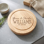 A round natural wood cutting board with engraved personalized text reading 'Grace & Charlie WILLIAMS EST. 21/09/2026', styled on a gray stone surface with a small bowl, burlap bag, and rosemary sprig nearby.