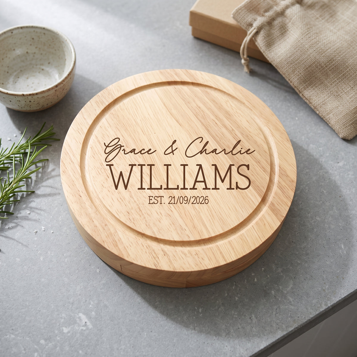 A round natural wood cutting board with engraved personalized text reading 'Grace & Charlie WILLIAMS EST. 21/09/2026', styled on a gray stone surface with a small bowl, burlap bag, and rosemary sprig nearby.