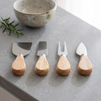 Four cheese and carving utensils with light wood handles and stainless steel heads are arranged on a gray stone surface, with a ceramic bowl and rosemary sprig in the background.