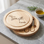 Personalised wooden cheese board set with a round lid engraved 'The Smith Family' and two matching round boards with cheese knives, styled on a gray marble surface with a small bowl of olive oil and rosemary.