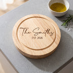 A round natural wood serving board engraved with 'The Smiths EST 2026' in dark script and block lettering, placed on a gray stone surface with a small bowl of dressing and rosemary nearby.