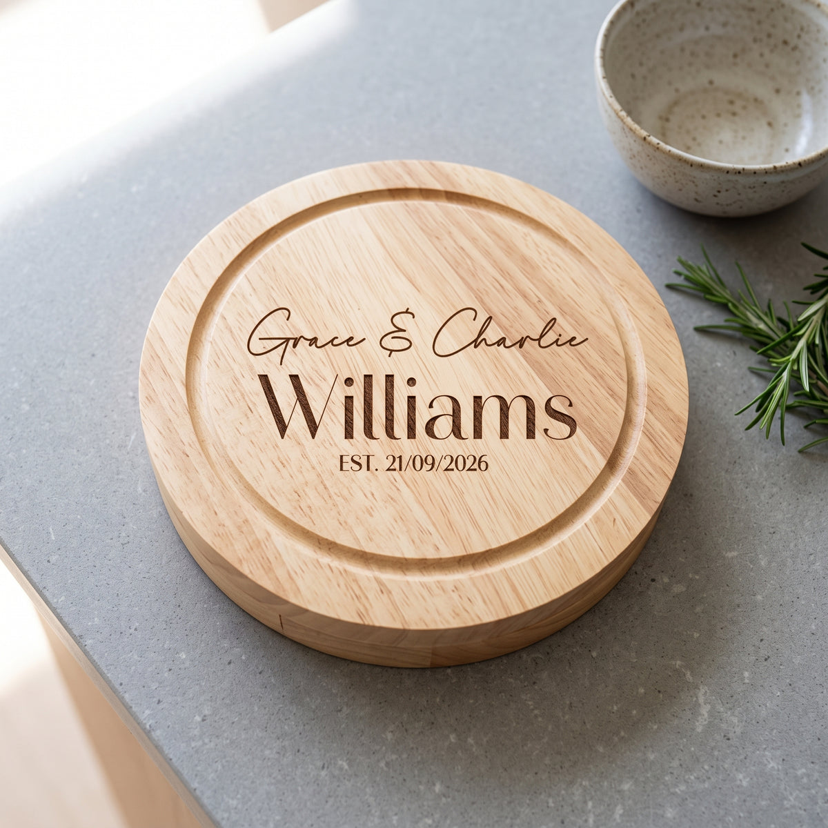 A round wooden cutting board with engraved names 'Grace & Charlie Williams' and a date, styled on a gray countertop with a ceramic bowl and rosemary sprigs.