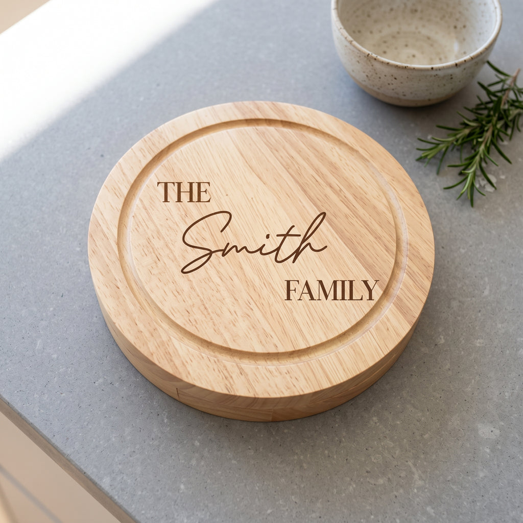 A round wooden serving board engraved with 'THE Smith FAMILY' in dark brown lettering, placed on a gray countertop with a small ceramic bowl and rosemary sprig nearby.