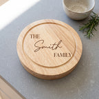 A round wooden serving board engraved with 'THE Smith FAMILY' in dark brown lettering, placed on a gray countertop with a small ceramic bowl and rosemary sprig nearby.