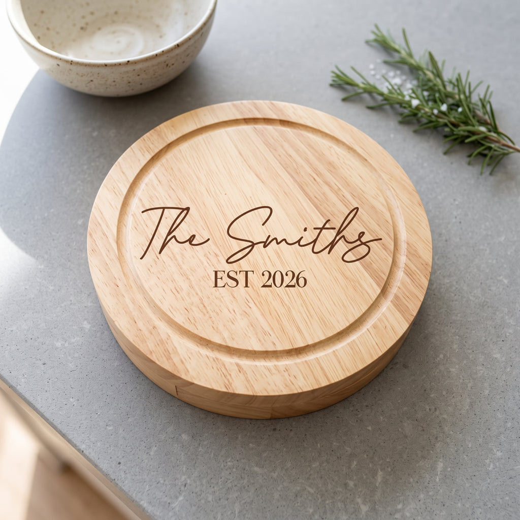 A round natural wood cutting board with engraved text reading 'The Smiths EST 2026', placed on a gray countertop with a ceramic bowl and rosemary sprig nearby.