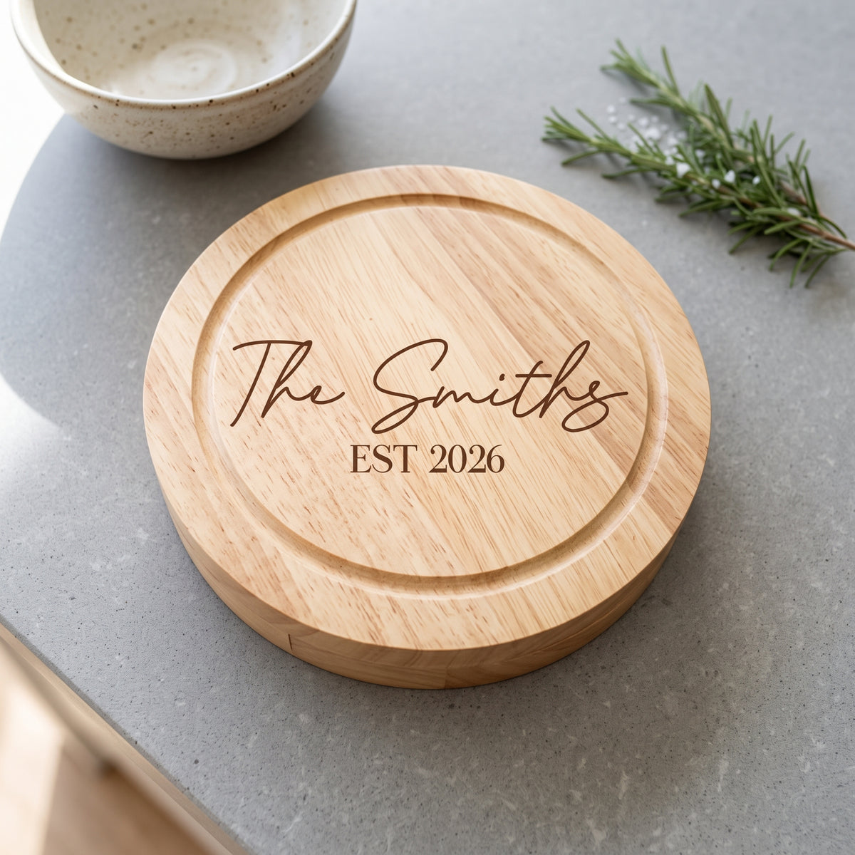 A round natural wood cutting board with engraved text reading 'The Smiths EST 2026', placed on a gray countertop with a ceramic bowl and rosemary sprig nearby.