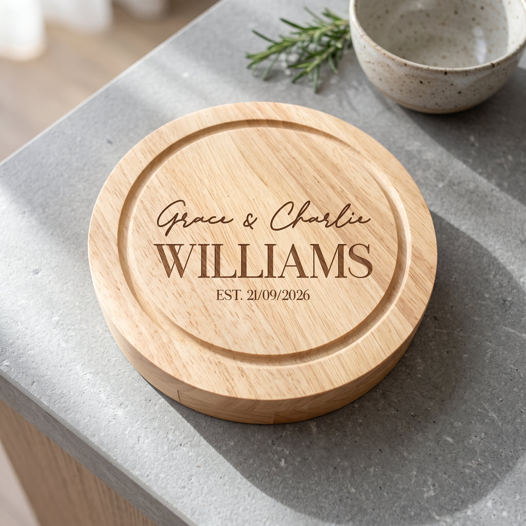 A round natural wood cutting board with personalized engraving reading 'Grace & Charlie WILLIAMS EST. 21/09/2026', placed on a gray stone countertop with a ceramic bowl and rosemary in the background.