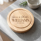 A round natural wood cutting board with personalized engraving reading 'Grace & Charlie WILLIAMS EST. 21/09/2026', placed on a gray stone countertop with a ceramic bowl and rosemary in the background.