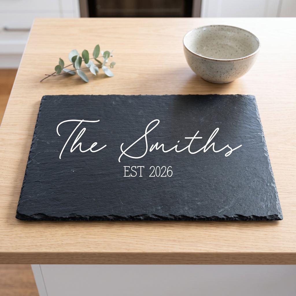 A personalised dark slate serving board with white script reading 'The Smiths' and 'EST 2026', placed on a light wood table with a ceramic bowl and eucalyptus sprig nearby.