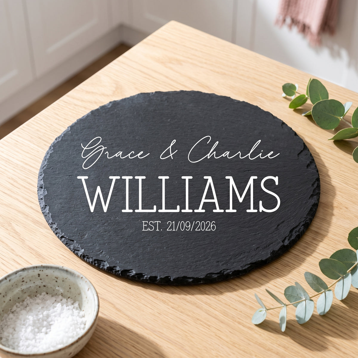 A round black slate serving board with white personalised text reading 'Grace & Charlie WILLIAMS EST. 21/09/2026', placed on a light wood table with eucalyptus leaves and a small bowl nearby.