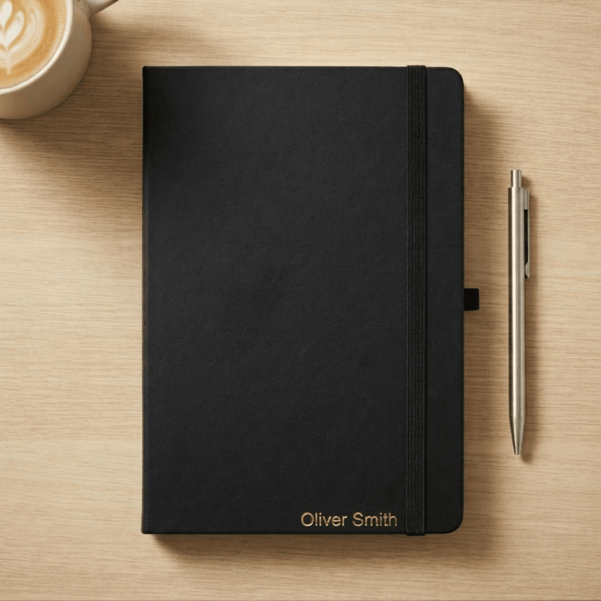 A black hardcover notebook with an elastic closure band and personalized gold text reading 'Oliver Smith' at the bottom, placed on a light wood tabletop next to a silver pen and a cup of coffee.