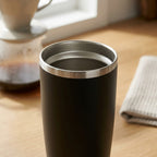 A matte black insulated travel tumbler with a brushed stainless steel rim, shown on a wooden table in a warm, softly lit setting with coffee gear blurred in the background.