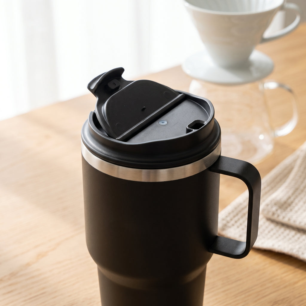 A matte black insulated travel tumbler with a carry handle and flip-top lid, shown on a wooden tabletop with a blurred coffee maker in the background.