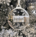 A personalised round silver metal Christmas ornament hanging on a frosted Christmas tree. The ornament features laser-cut snowflake designs at the top and bottom and a central banner with the name 'Jordan' in black text.