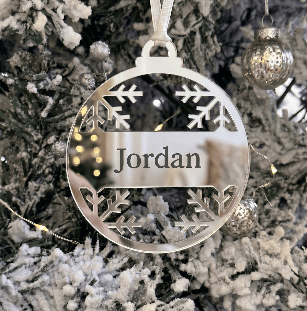 A personalised round silver metal Christmas ornament hanging on a frosted Christmas tree. The ornament features laser-cut snowflake designs at the top and bottom and a central banner with the name 'Jordan' in black text.