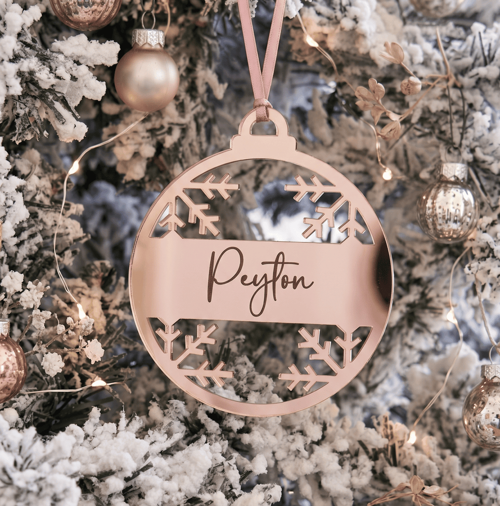 A personalised round acrylic Christmas ornament in a rose gold finish hangs from a pink ribbon on a snowy flocked Christmas tree. The center of the ornament features a horizontal bar with the name 'Peyton' engraved in elegant cursive, surrounded by cutout snowflake designs.