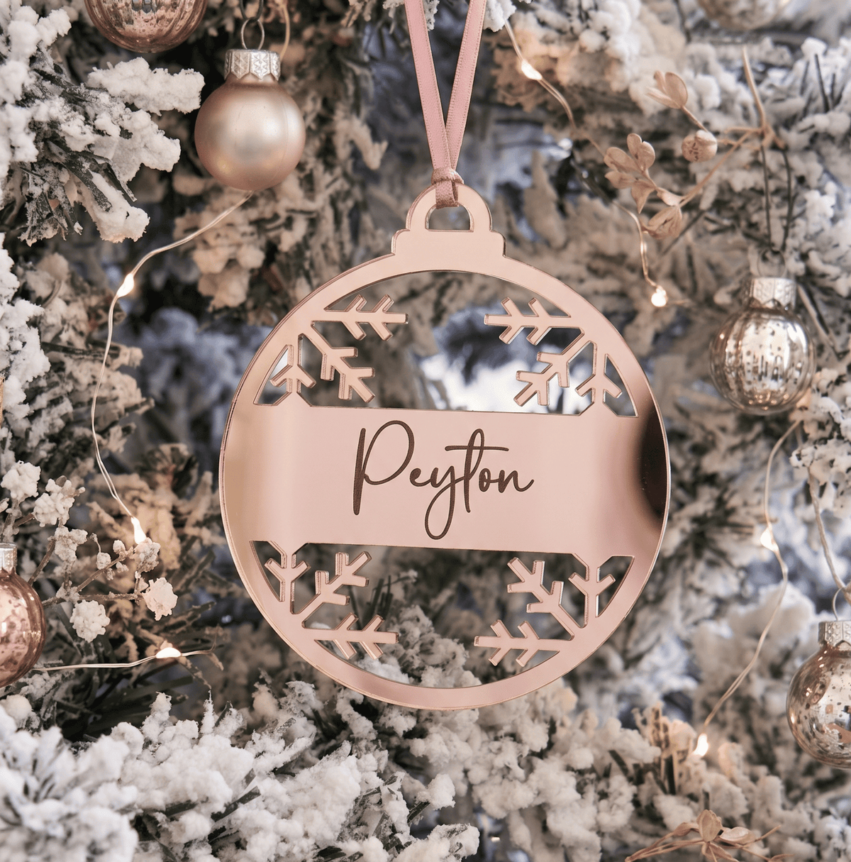 A personalised round acrylic Christmas ornament in a rose gold finish hangs from a pink ribbon on a snowy flocked Christmas tree. The center of the ornament features a horizontal bar with the name 'Peyton' engraved in elegant cursive, surrounded by cutout snowflake designs.