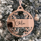A round rose gold acrylic Christmas ornament with cutout snowflakes and the name 'Chloe' in script, hanging on a decorated snowy Christmas tree with lights.