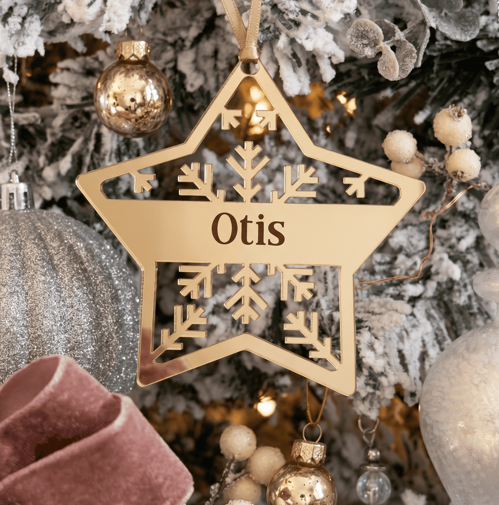 A personalised wooden star Christmas ornament engraved with the name 'Otis' and a snowflake design, hanging on a decorated Christmas tree with gold and silver ornaments.