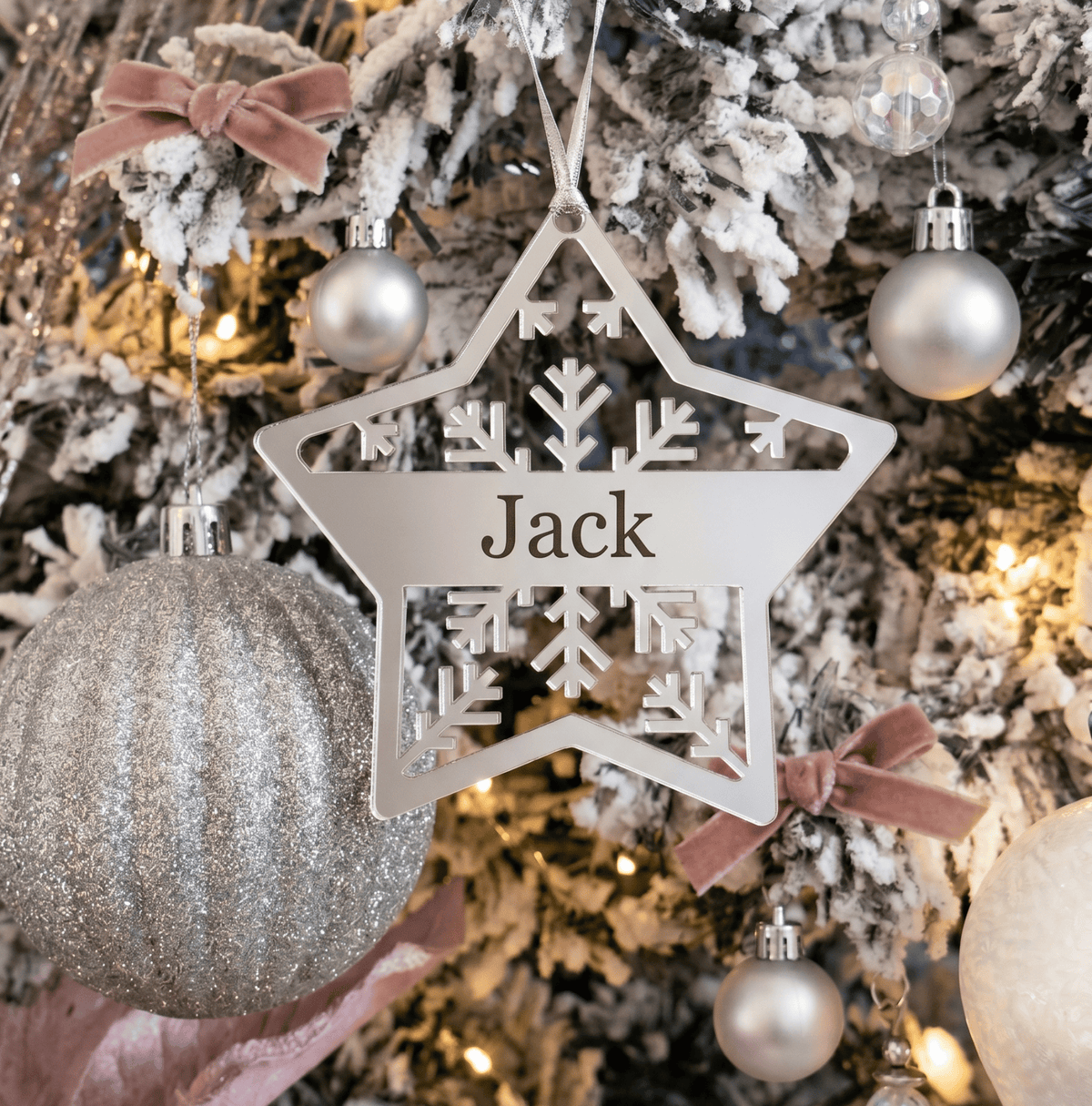 A personalised silver acrylic star Christmas ornament with the name 'Jack' engraved in the centre, hanging on a decorated flocked Christmas tree with silver ornaments and soft pink ribbons.