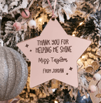 A personalised star-shaped Christmas ornament hanging on a decorated tree. The ornament is pale pink with dark brown text that reads 'THANK YOU FOR HELPING ME SHINE Miss Taylor FROM JORDAN'.