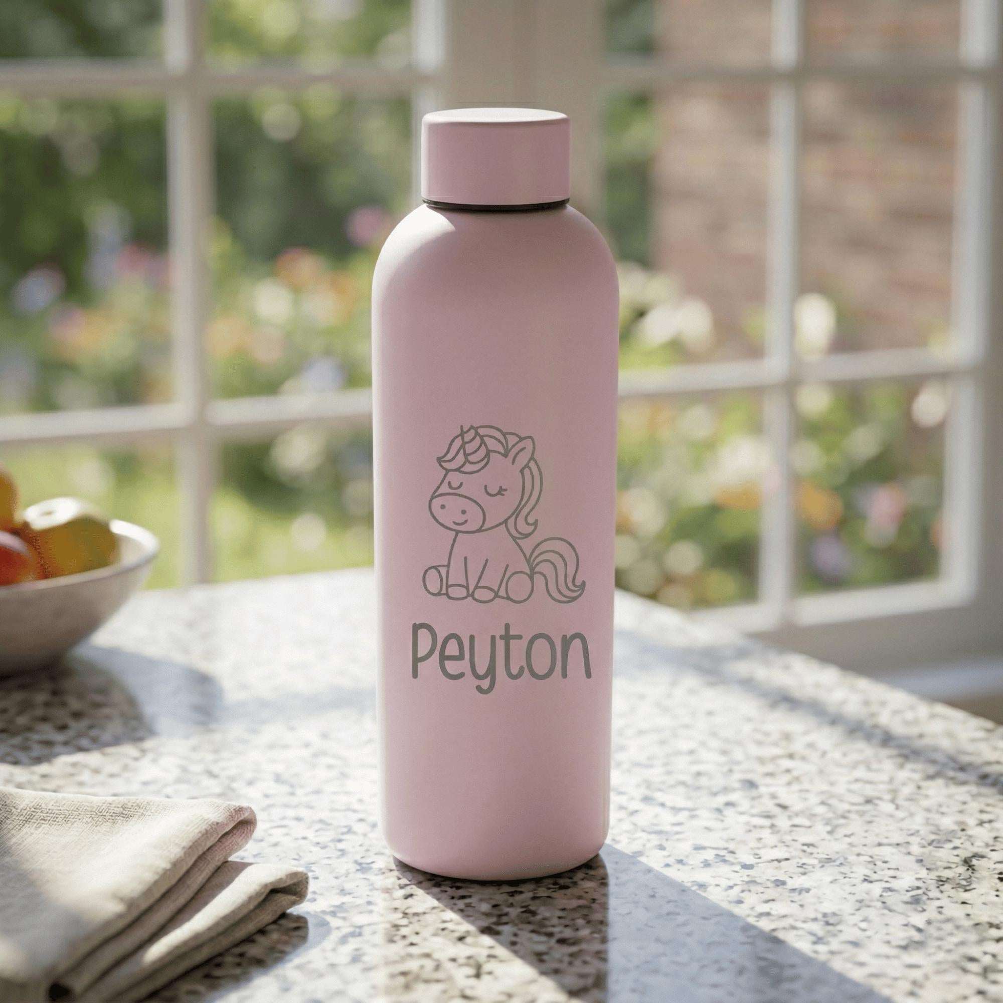 Pink water bottle with unicorn design and 'Peyton' text on a table with a blurred garden background