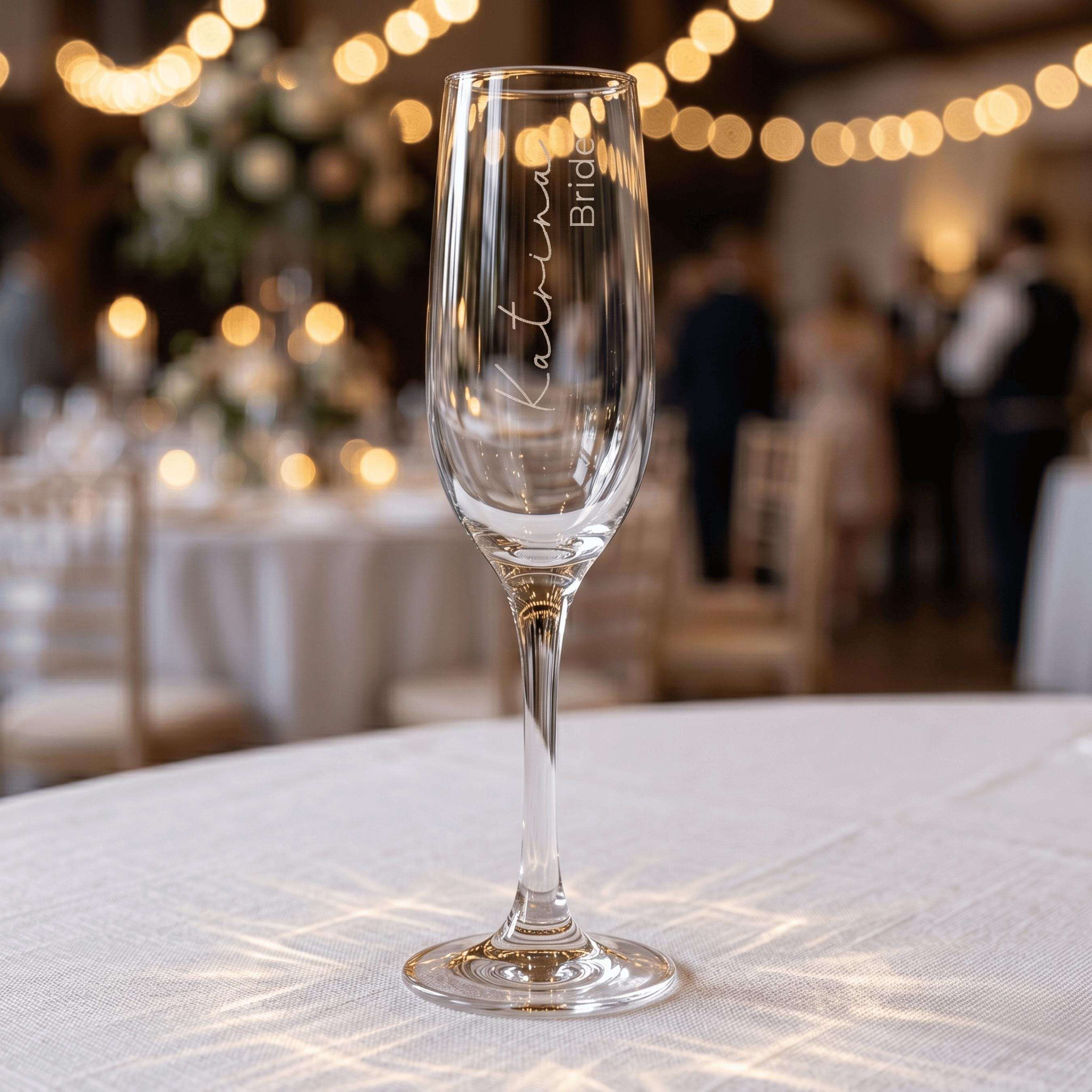 Personalised wedding champagne flute engraved with "Katrina Bride" in elegant script, standing on a white tablecloth with festive wedding venue lighting in the background.