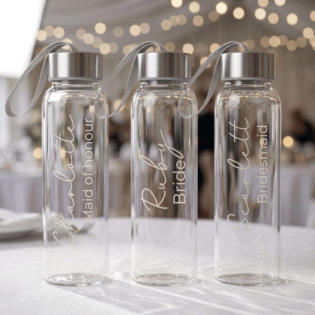Three personalised glass water bottles for a bridal party featuring "Charlotte Maid of honour", "Ruby Bride", and "Scarlett Bridesmaid" in elegant white script, displayed at a wedding reception.
