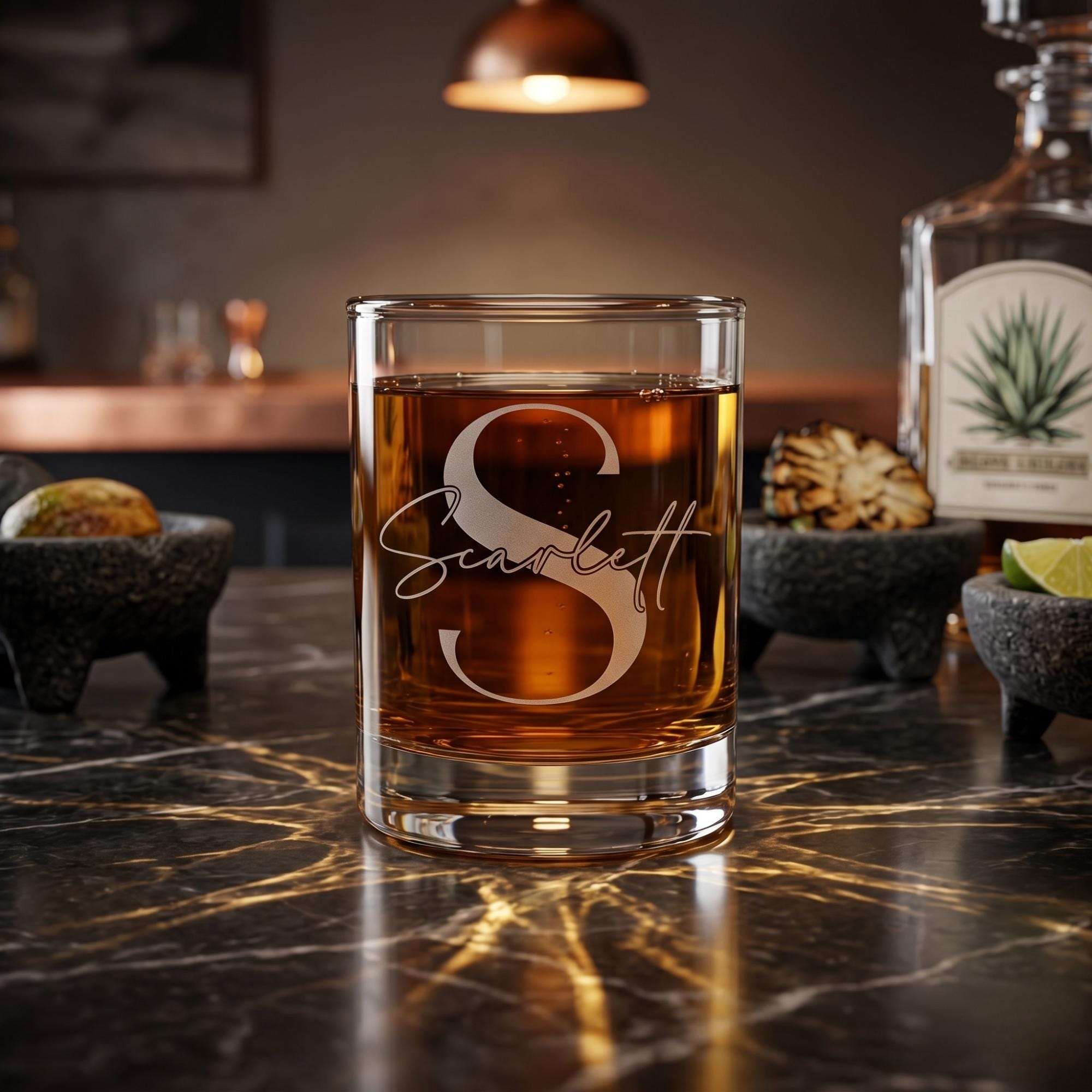 Glass of amber liquid with 'Scarlett' engraving on a dark marble surface, with a bottle and ingredients in the background.