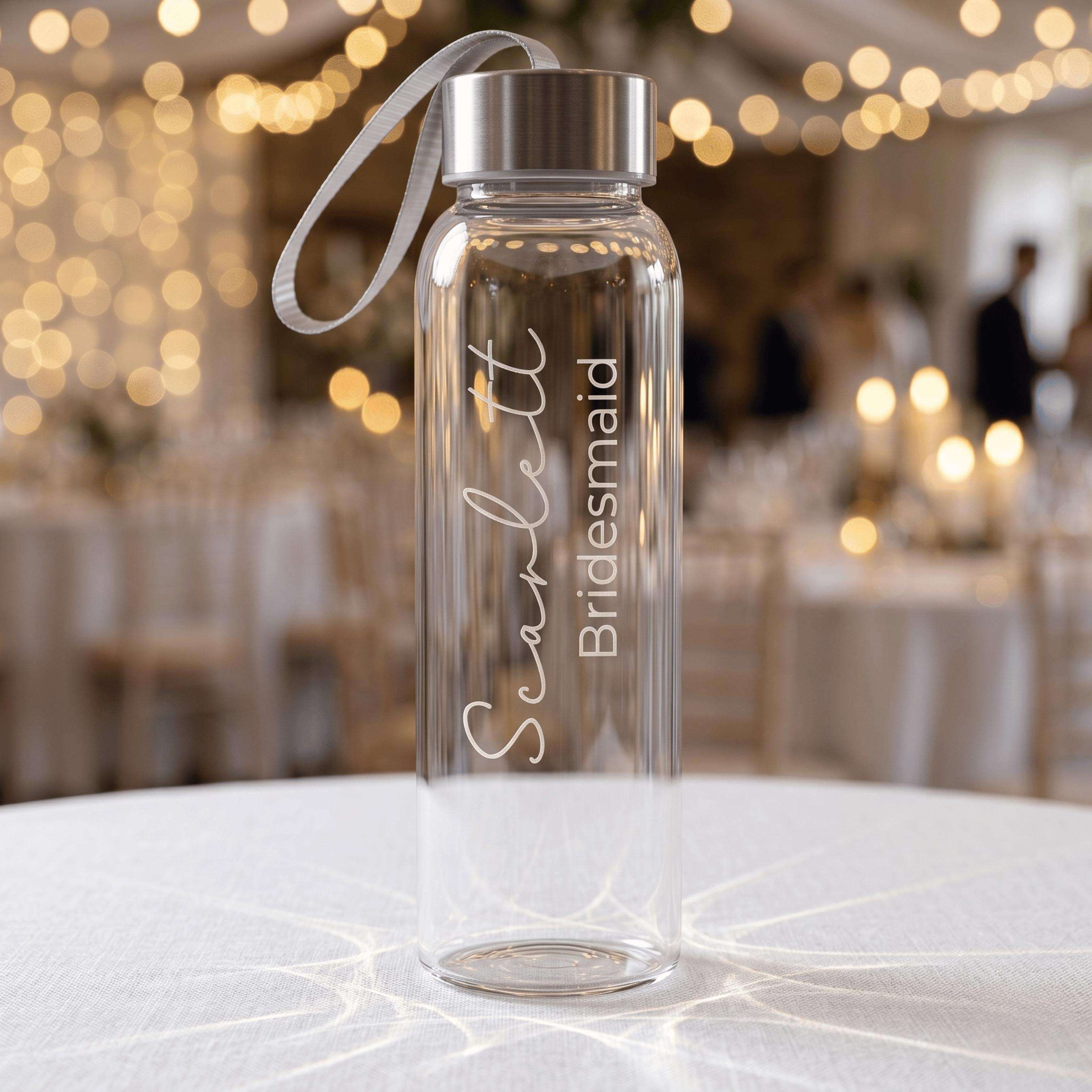 Personalised bridesmaid gift: a clear glass water bottle engraved with "Scarlett Bridesmaid" in a stylish script font, shown in a festive wedding venue setting with warm lighting.