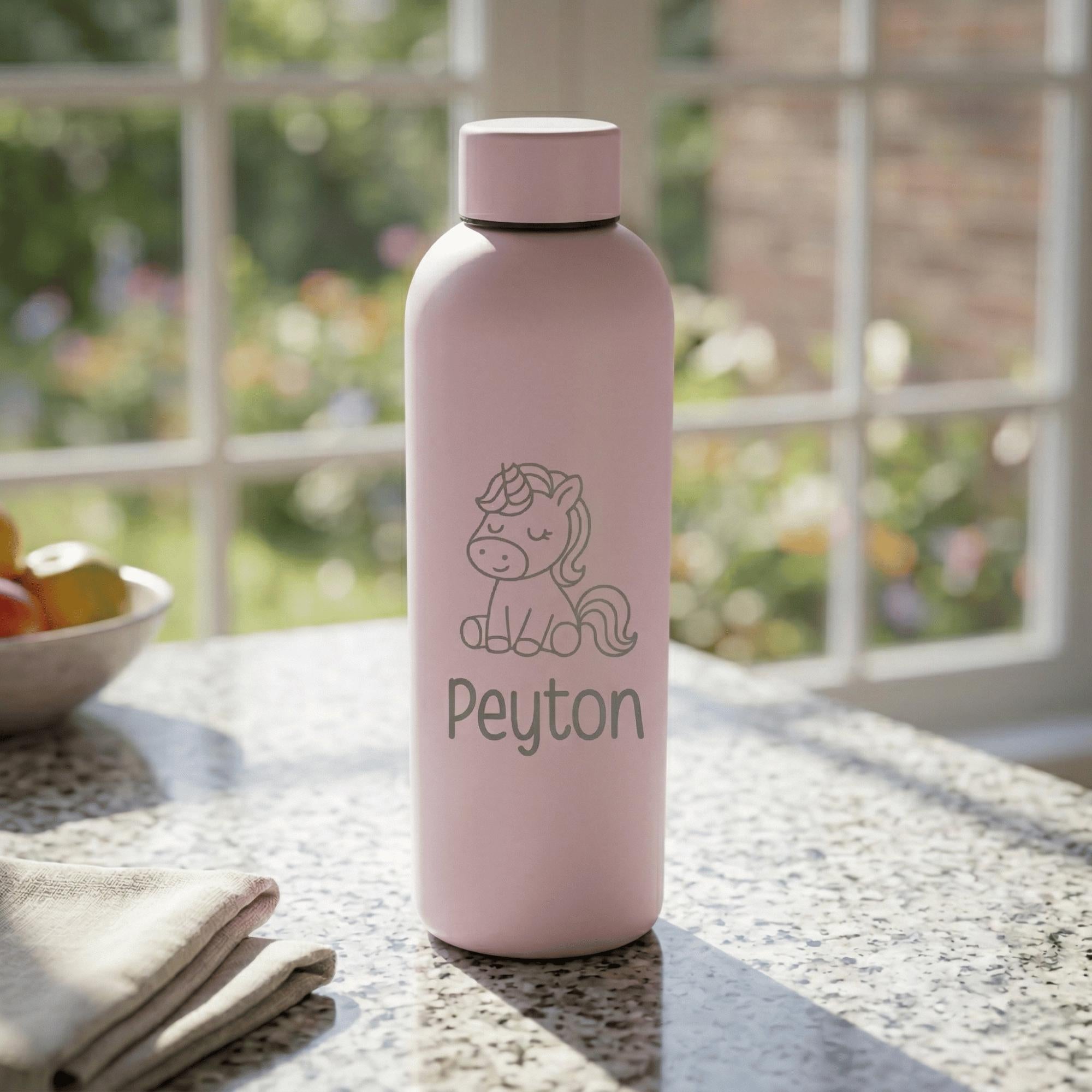 Pink water bottle with unicorn design and 'Peyton' text on a table with a blurred garden background
