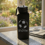 Black water bottle with soccer ball design and 'Andrew' text on a windowsill with garden view