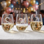 Three personalized wine glasses with names on a table with balloons and people in the background