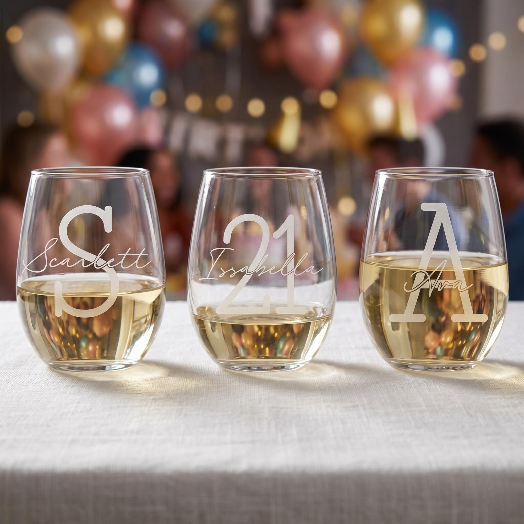 Three personalized wine glasses with names on a table with balloons and people in the background