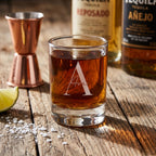 Glass of tequila with 'Ava' branding on a wooden surface with bottles and bar tools in the background.