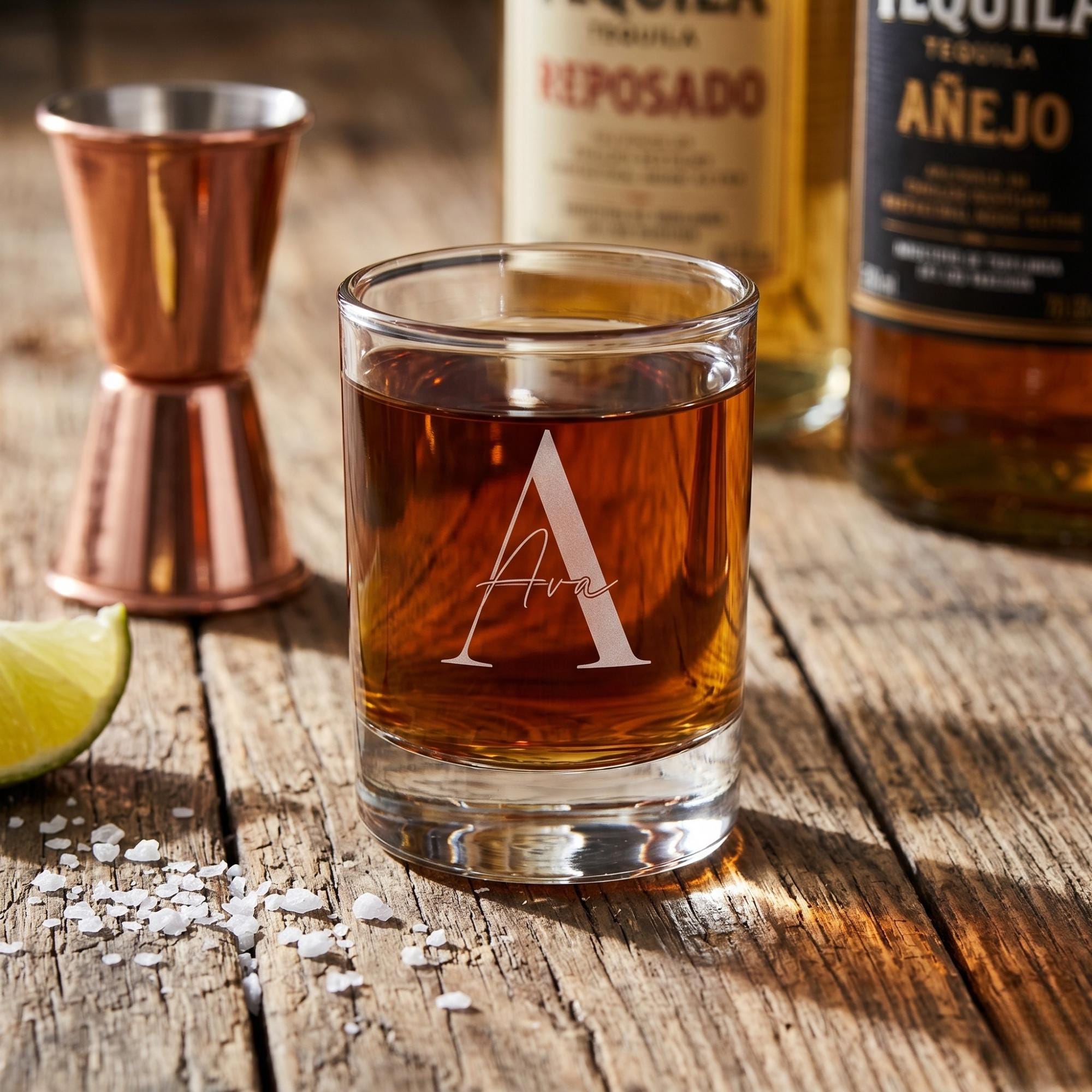 Glass of tequila with 'Ava' branding on a wooden surface with bottles and bar tools in the background.