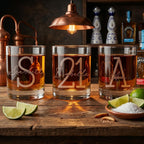Three glasses of whiskey with personalized engravings on a wooden table with limes and salt.