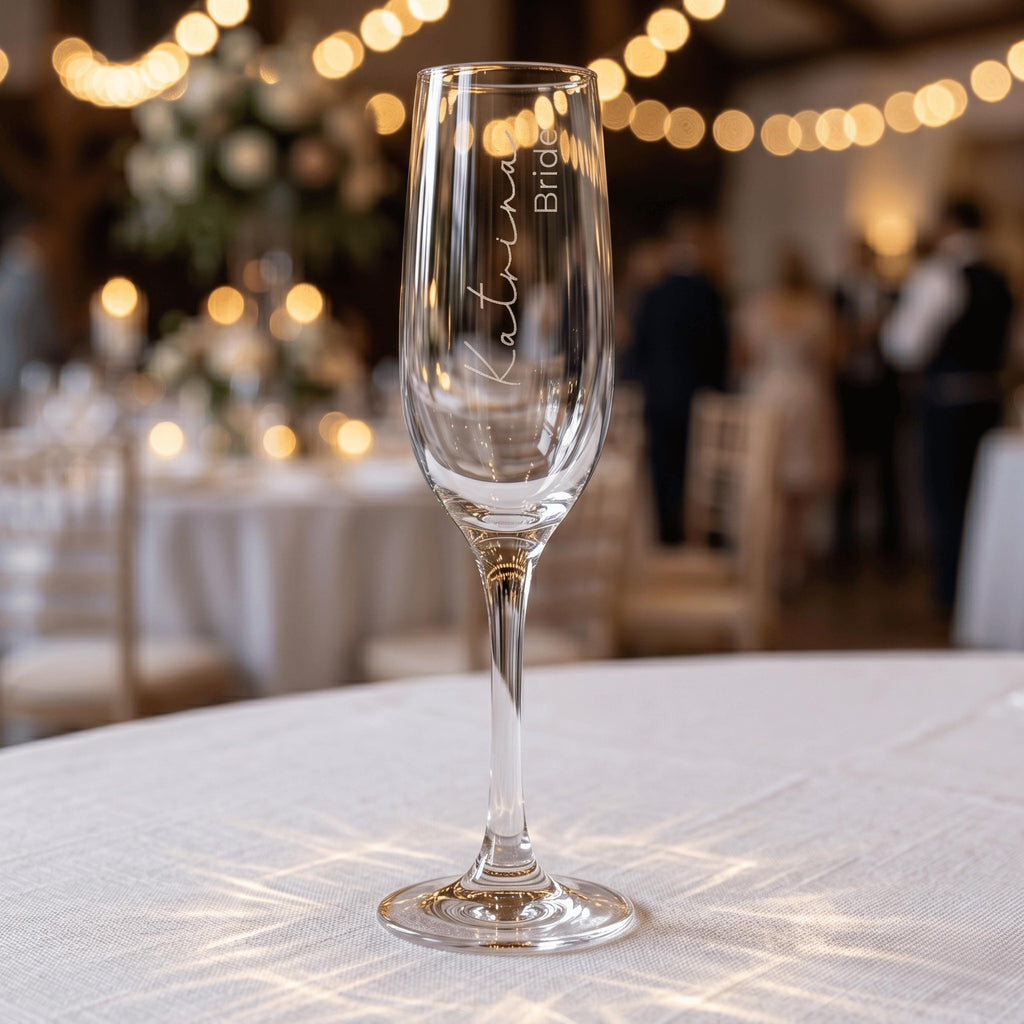 Personalised wedding champagne flute engraved with "Katrina Bride" in elegant script, standing on a white tablecloth with festive wedding venue lighting in the background.
