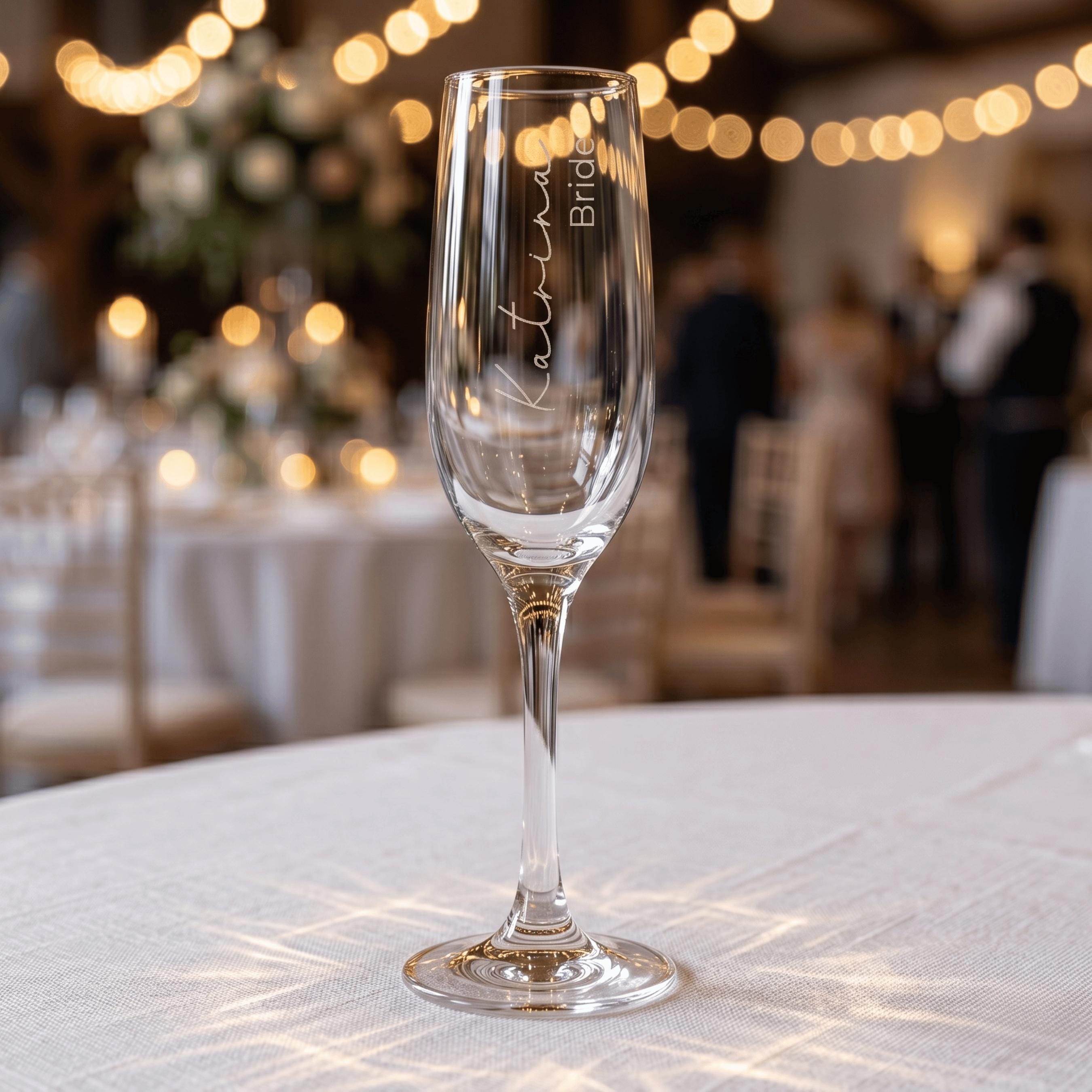 Personalised wedding champagne flute engraved with "Katrina Bride" in elegant script, standing on a white tablecloth with festive wedding venue lighting in the background.