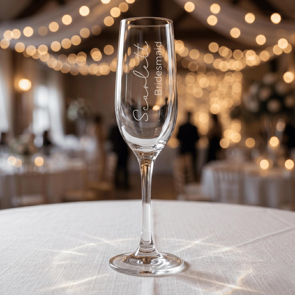 Personalised bridesmaid gift champagne flute with "Scarlett Bridesmaid" engraving, displayed on a table at a wedding celebration with elegant string lights.